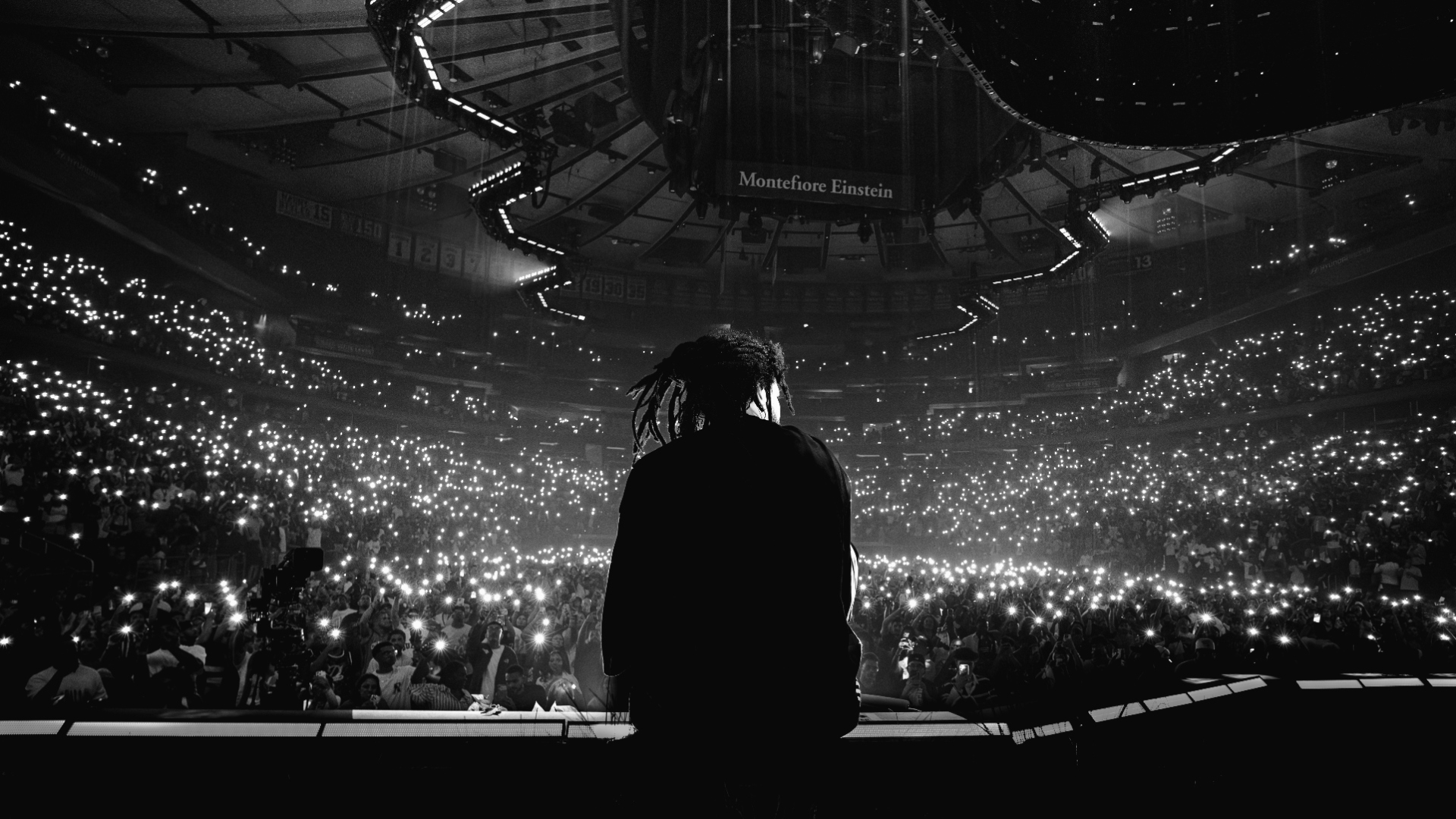 J. Cole silhouette on stage at Madison Square Garden, phone lights across the arena